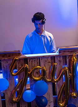 In einem festlich dekorierten Raum mixt ein DJ, während goldene Buchstaben und blaue Ballons die Stimmung heben.