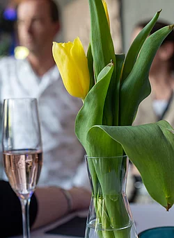 Eine frische gelbe Tulpe in einer transparenten Vase steht im Vordergrund, während im Hintergrund unscharf Menschen zu sehen sind.