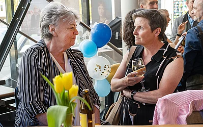 In einem festlichen Ambiente stehen zwei Frauen am Tisch, umgeben von Luftballons und einer Vase mit gelben Tulpen.