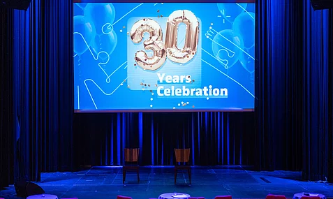 Eine festliche Bühne mit blauen Vorhängen, auf der ein großes Banner mit der Aufschrift '30 Jahre Feier' prangt.